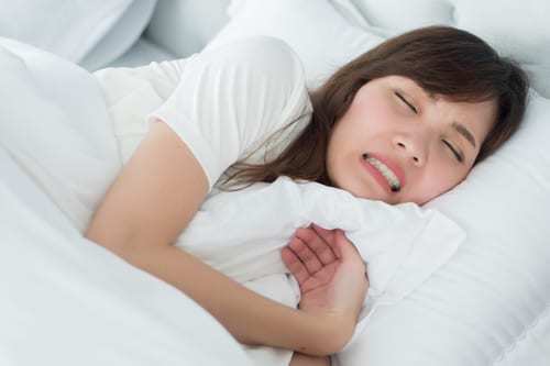 Woman laying in bed with white sheets grinding her teeth