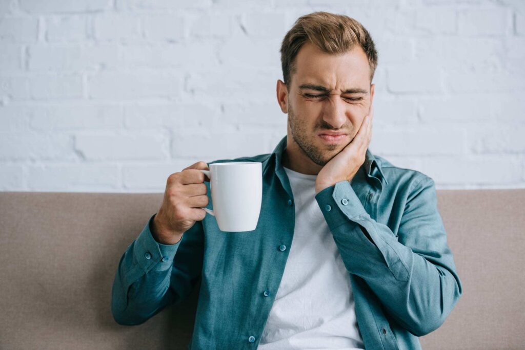 Man holding side of face near need, grimacing in pain while holding coffee cup in other hand