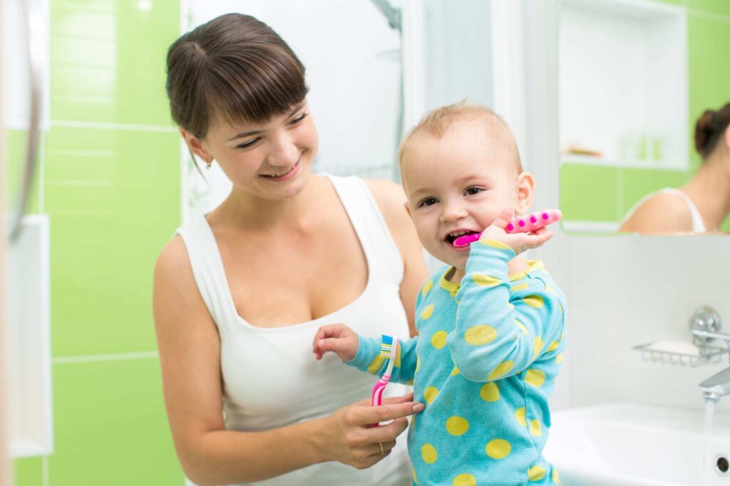 Mother and baby brushing teeth in bathroom with green tile.